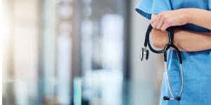 Close-up of a healthcare professional wearing blue scrubs, holding a stethoscope in crossed arms. The background is blurred, suggesting a modern, well-lit medical facility or hospital setting.